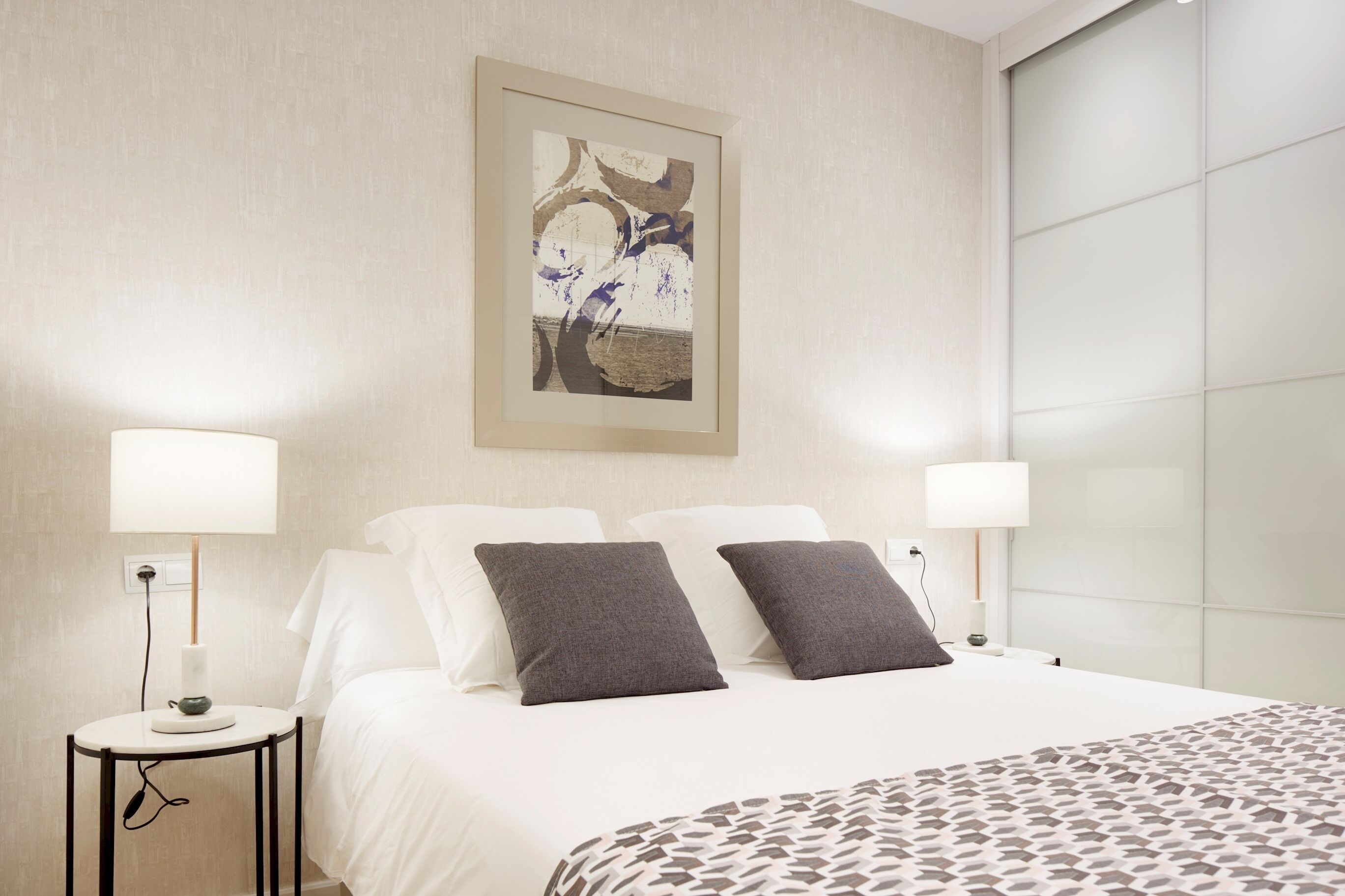 A hotel room with a bed, two bedside tables with lamps, a framed artwork on the wall, and a large window.