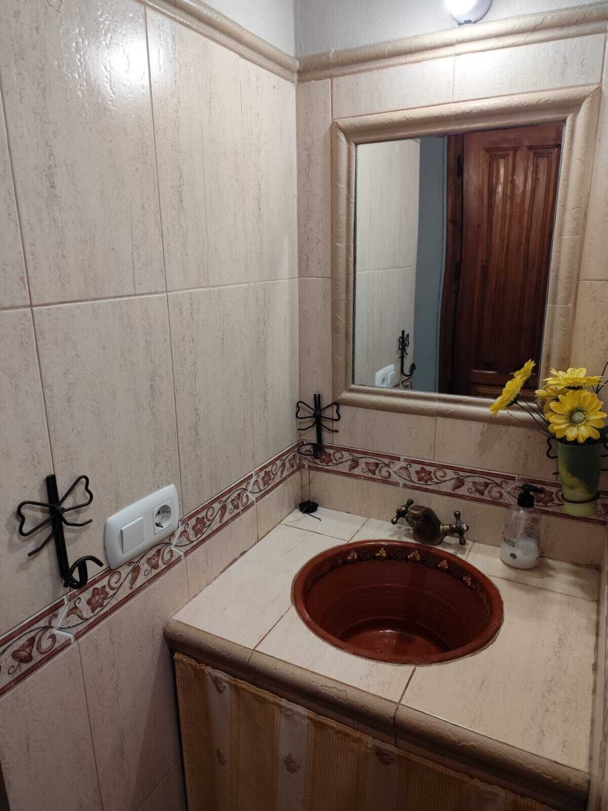 A bathroom with a tiled wall, a wooden door, a mirror, a sink with a red basin, a towel rack, and a vase with yellow flowers.