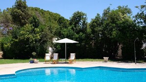 A swimming pool with clear blue water, surrounded by a concrete deck. There are three lounge chairs and a white umbrella providing shade. The area is bordered by lush green trees and a hill in the background. - Villa with 7 rooms, swimming pool, tennis court and garden (Alvor)