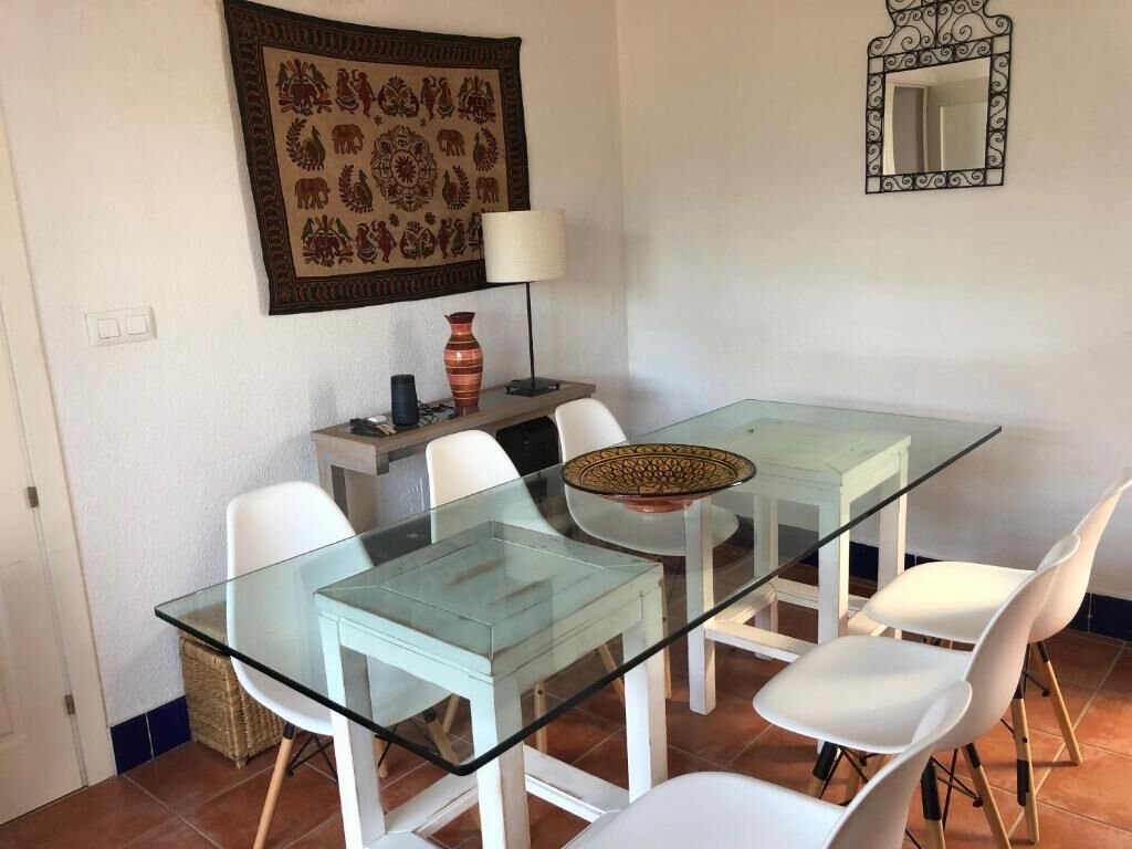 A dining area with a glass-top table, white chairs, a decorative bowl, a vase, a lamp, and a wall tapestry.