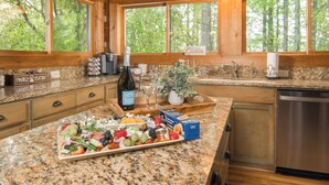 A kitchen with granite countertops, wooden cabinets, a built-in dishwasher, and a large window overlooking a forest. - Mountain Serenity! King Beds - Hot Tub - Fireplace - Ping Pong - Fire Pit (Boone)