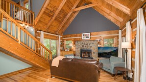 A spacious living area with a stone fireplace, wooden staircase, and a cozy couch. - Mountain Serenity! King Beds - Hot Tub - Fireplace - Ping Pong - Fire Pit (Boone)