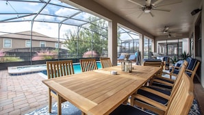 A covered patio with a wooden table and chairs, a pool, and a view of a house in the background. - Luxury Champions Gate Pool Home | Upgrades, Game Room, Spa & BBQ (Champions Gate)