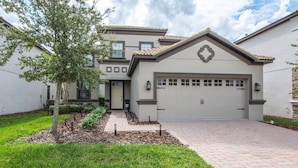 A two-story house with a garage, a landscaped front yard, and a brick driveway. - Luxury Champions Gate Pool Home | Upgrades, Game Room, Spa & BBQ (Champions Gate)