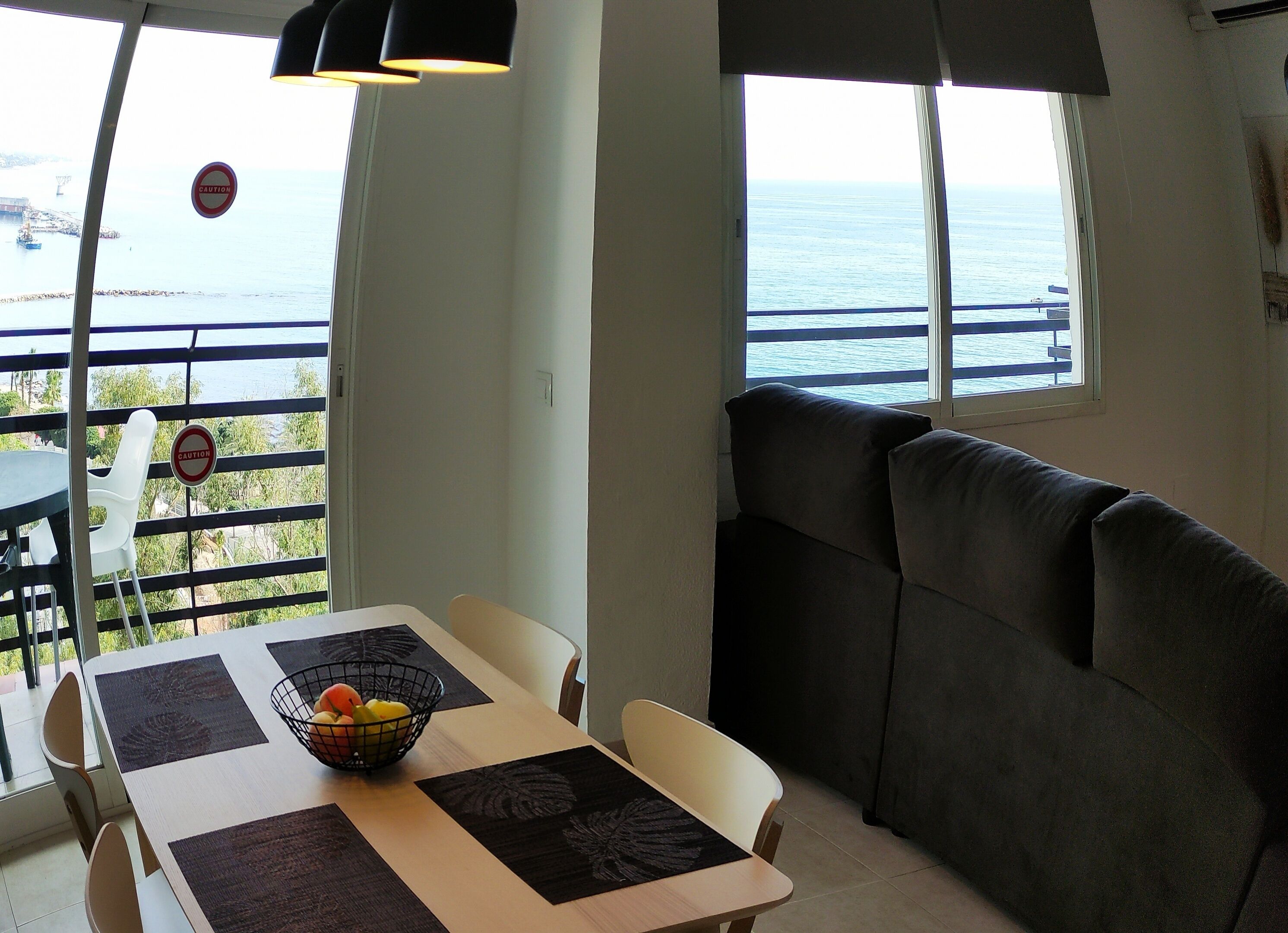 Uma área de refeições com uma mesa posta para dois, um banco acolchoado e vista para o mar através de grandes janelas.
