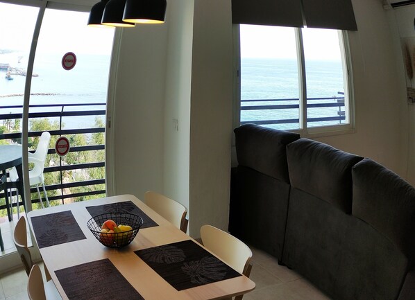 A dining area with a table set for two, a cushioned bench, and a view of the sea through large windows.