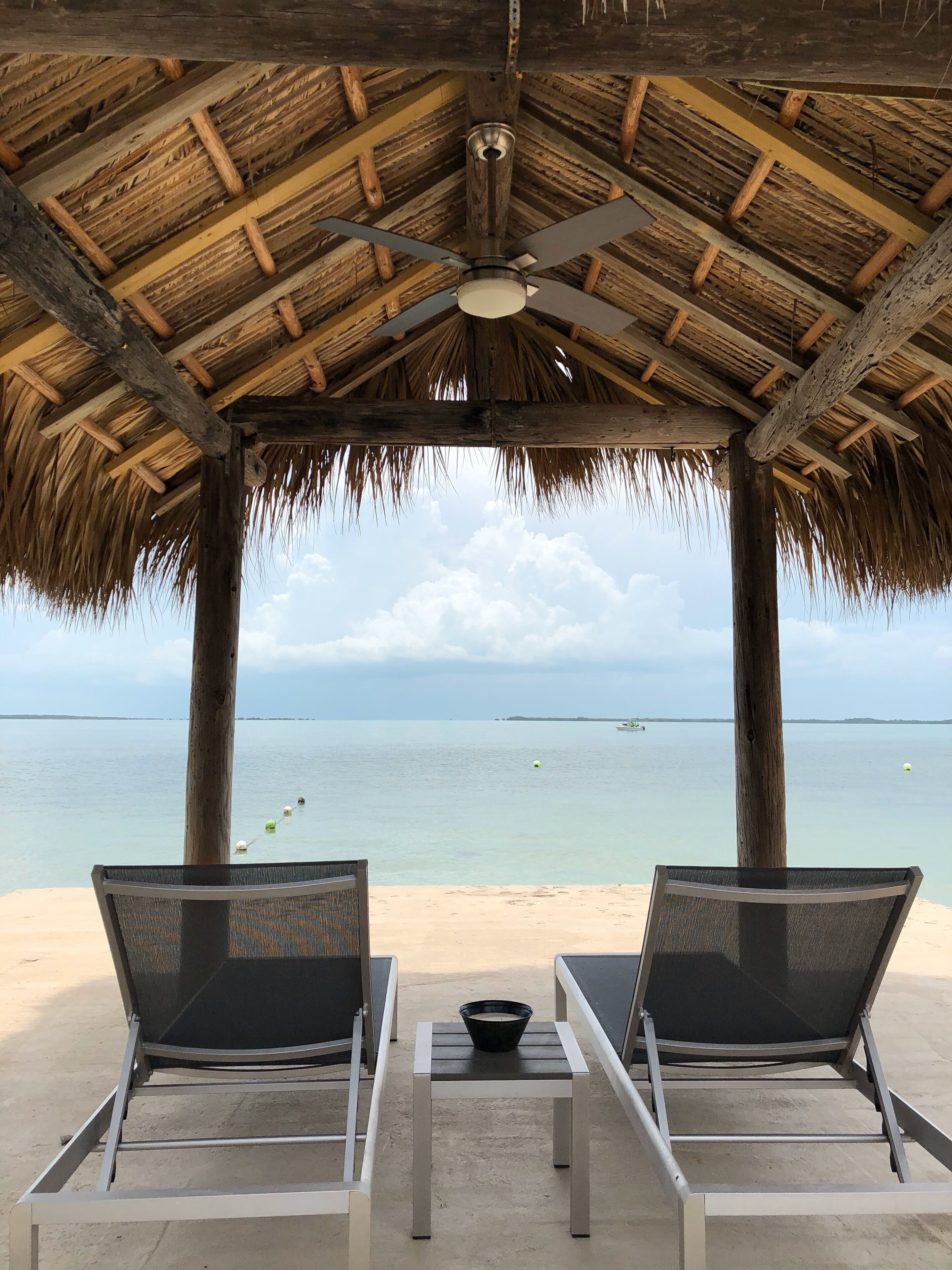 A beachside gazebo with a thatched roof, two lounge chairs, and a table with a bowl.