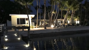 A wooden dock leading to a modern building with a pool, surrounded by palm trees and illuminated by string lights. - Sunset Villas: Bayfront Beach Compound<br>**28 Night Min** (Key Largo)