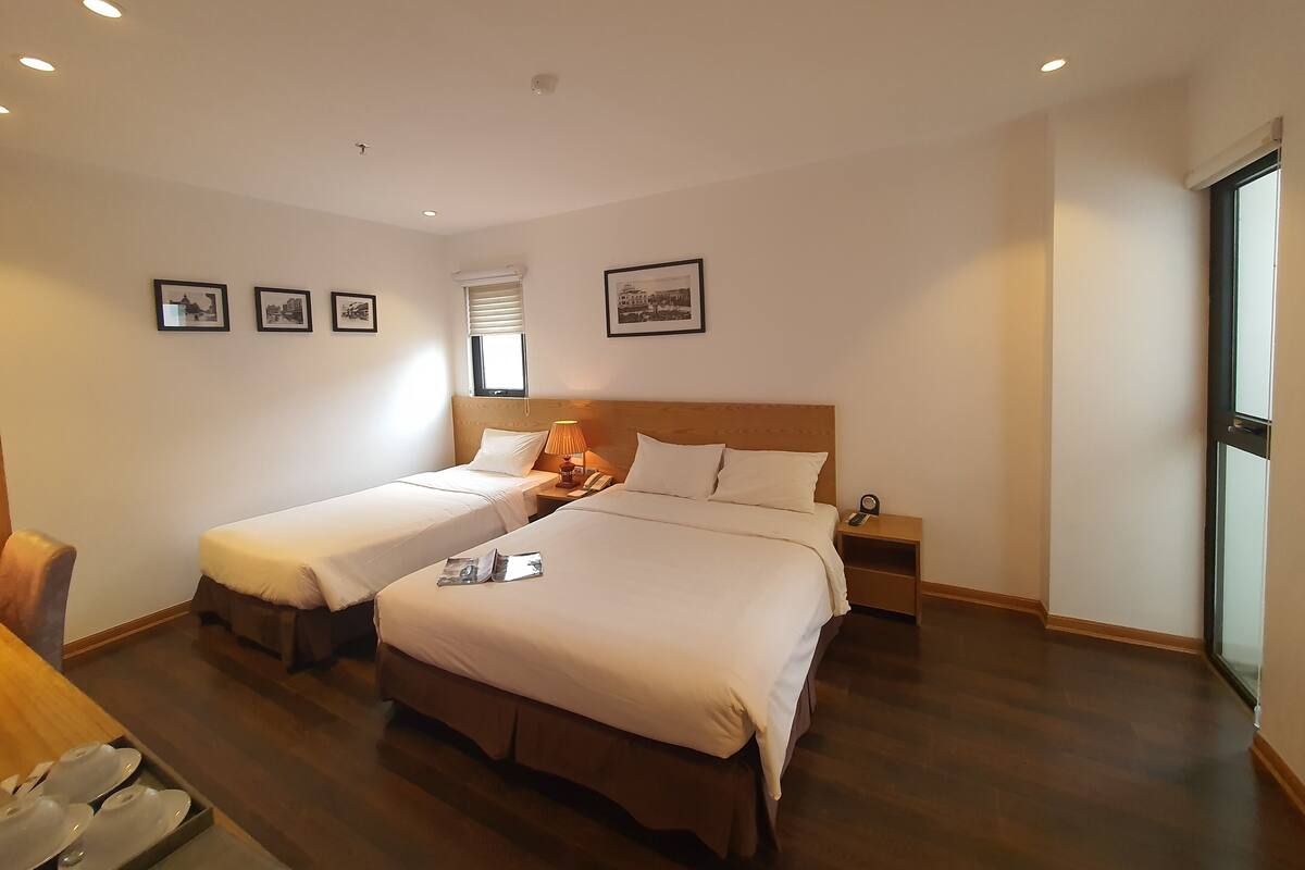 Una habitación de hotel con dos camas individuales, un cabecero de madera, una mesita de noche con una lámpara, una ventana con persianas y cuadros enmarcados en la pared.