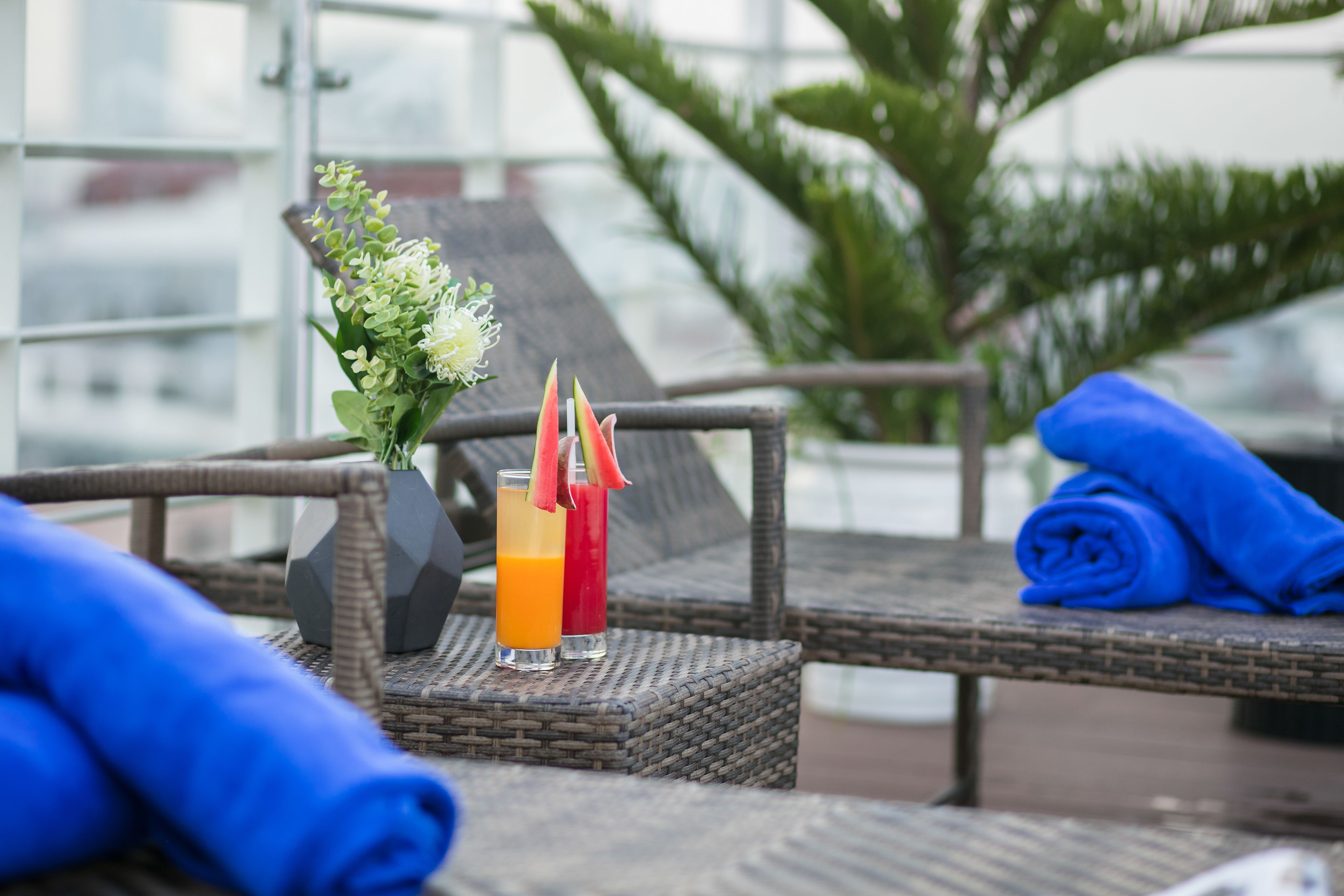 A wicker lounge chair with a blue towel, two drinks with fruit garnishes, and a small vase with greenery.
