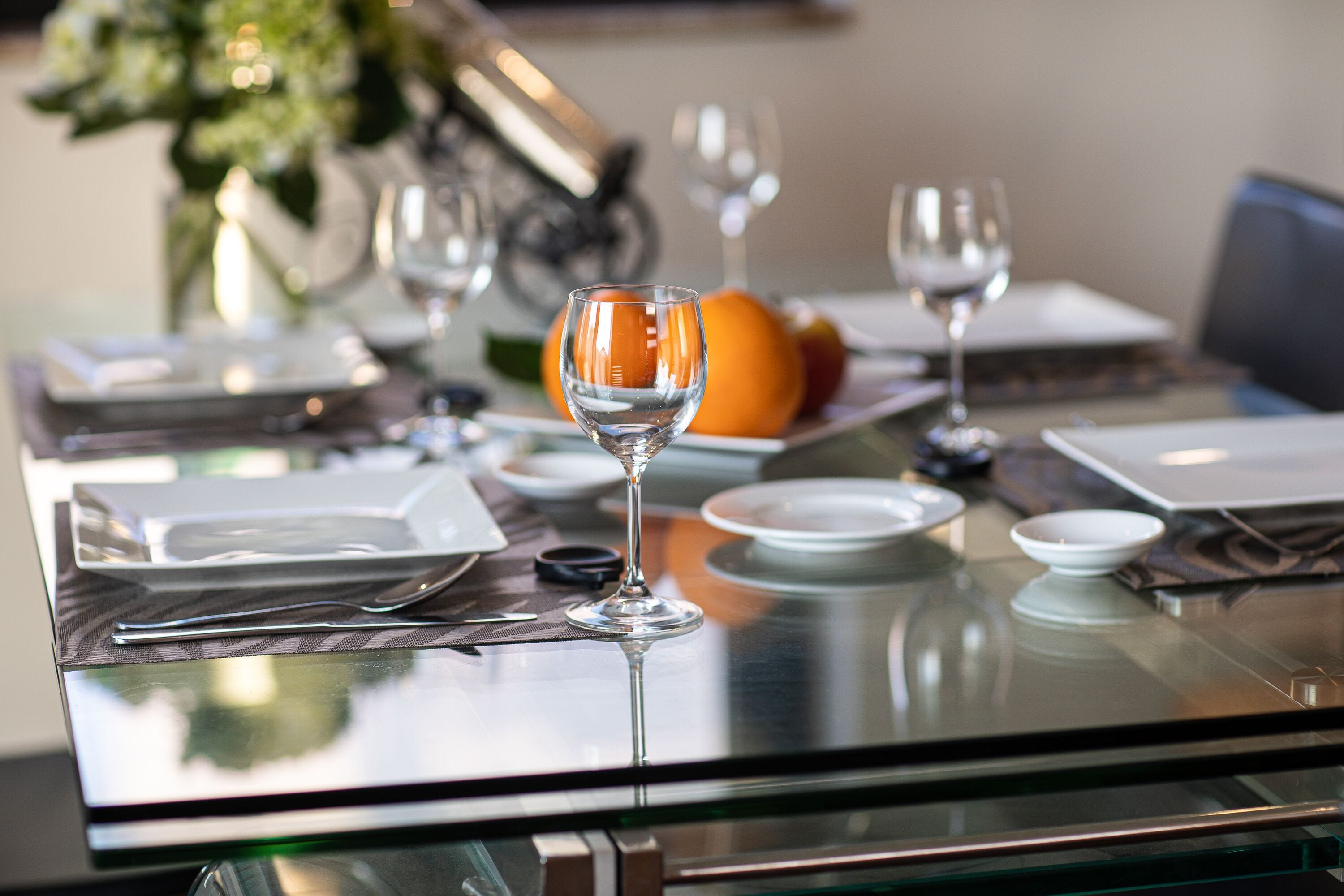 Una mesa de comedor servida con platos, cubiertos y copas de vino, con un frutero y un florero al fondo.