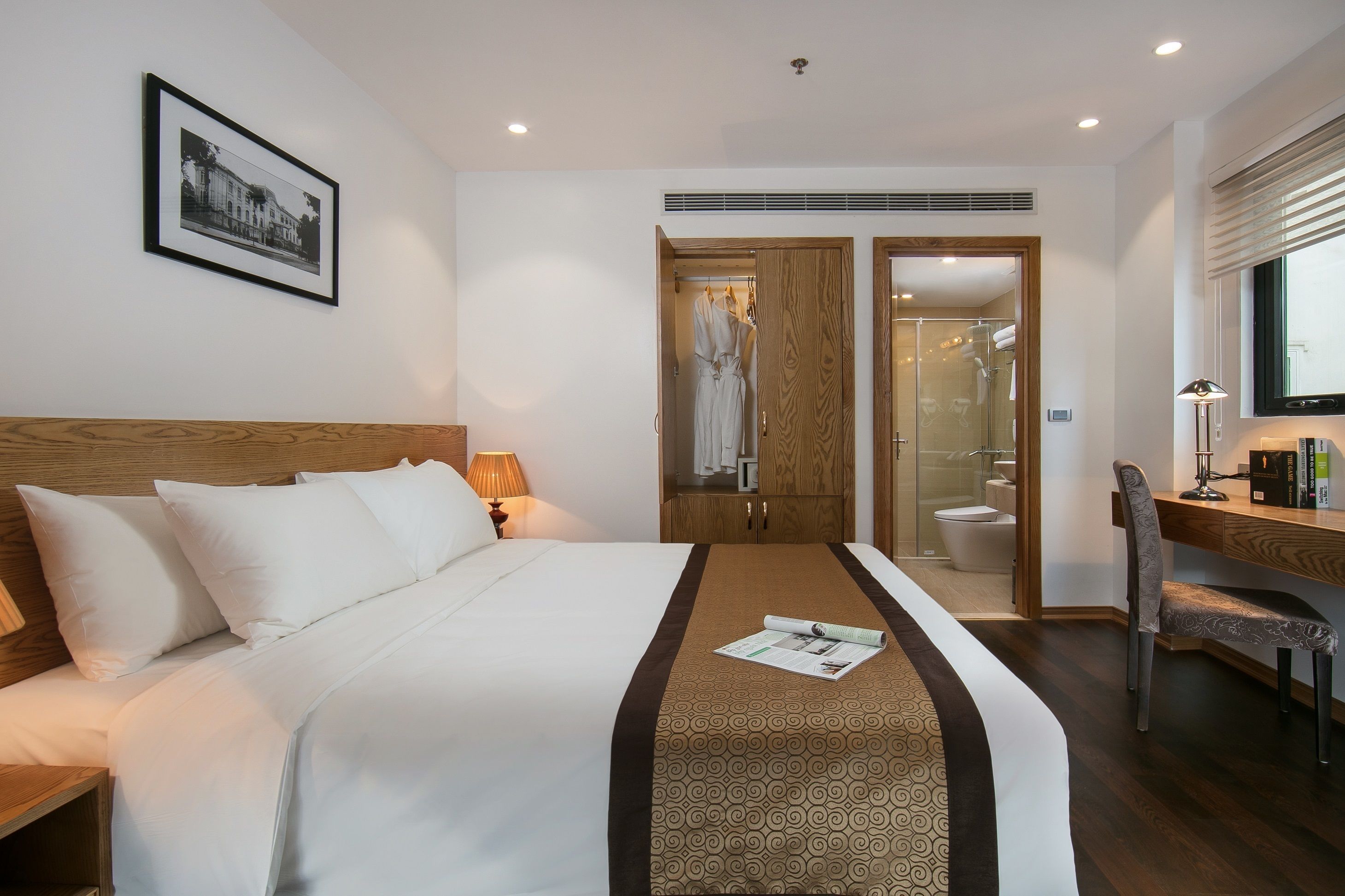 A hotel room with a neatly made bed, a desk with a chair, a shower area, and a framed picture on the wall.