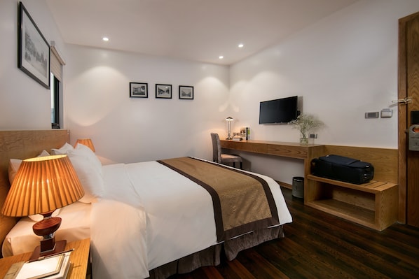 Una habitación de hotel con cama, mesita de noche, lámpara, escritorio, silla, televisor y una planta pequeña.