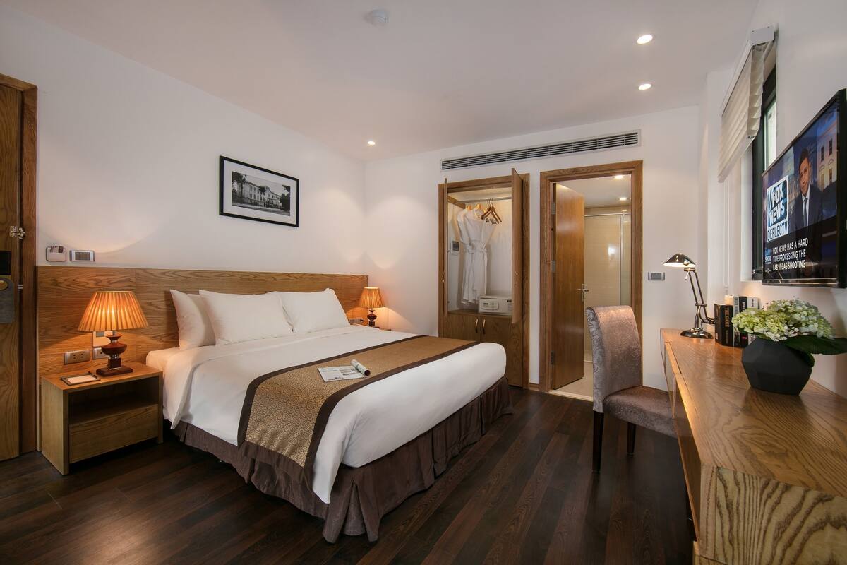 Una habitación de hotel con una cama grande, mesitas de noche, un escritorio con silla, televisor y espejo.