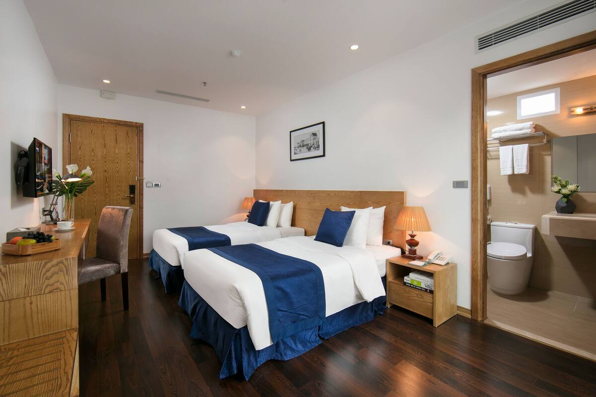 Habitación de hotel con dos camas, un escritorio, una silla, una mesita de noche con lámpara, un cuadro en la pared y un baño con inodoro y lavamanos.