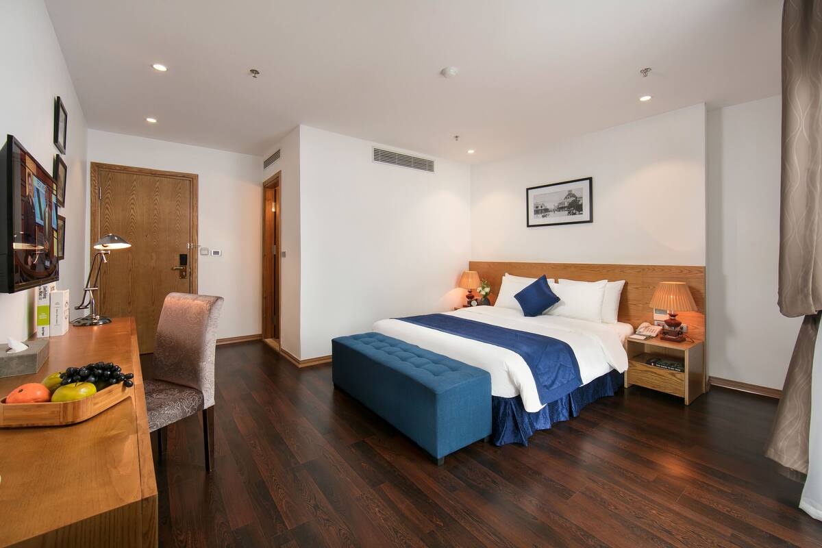 Habitación de hotel con piso de madera, cama con ropa de cama azul, escritorio con frutero y una silla.