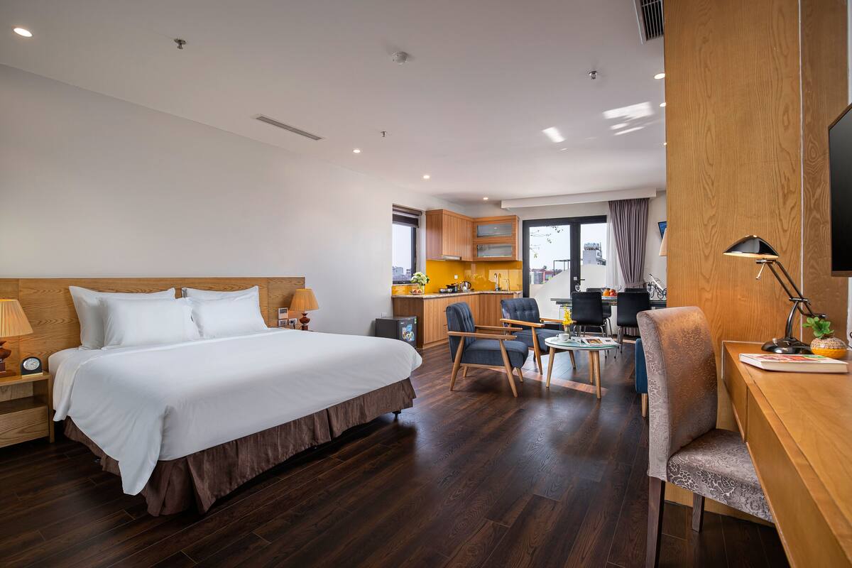Una habitación de hotel con una cama grande, un escritorio con una silla, una mesita pequeña y una kitchenette al fondo.