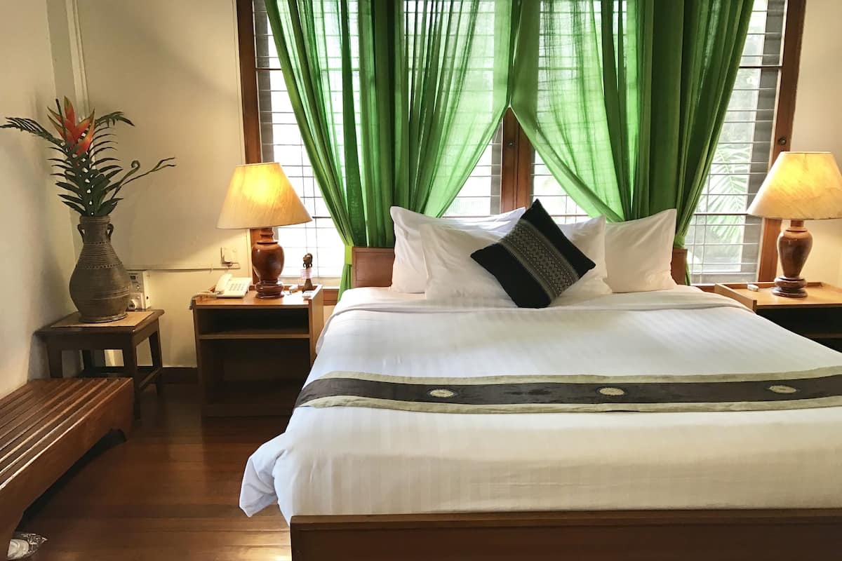 A neatly made bed with white linens and a black pillow with a geometric pattern. There are two bedside lamps on wooden nightstands, and a potted plant on the left side of the bed.