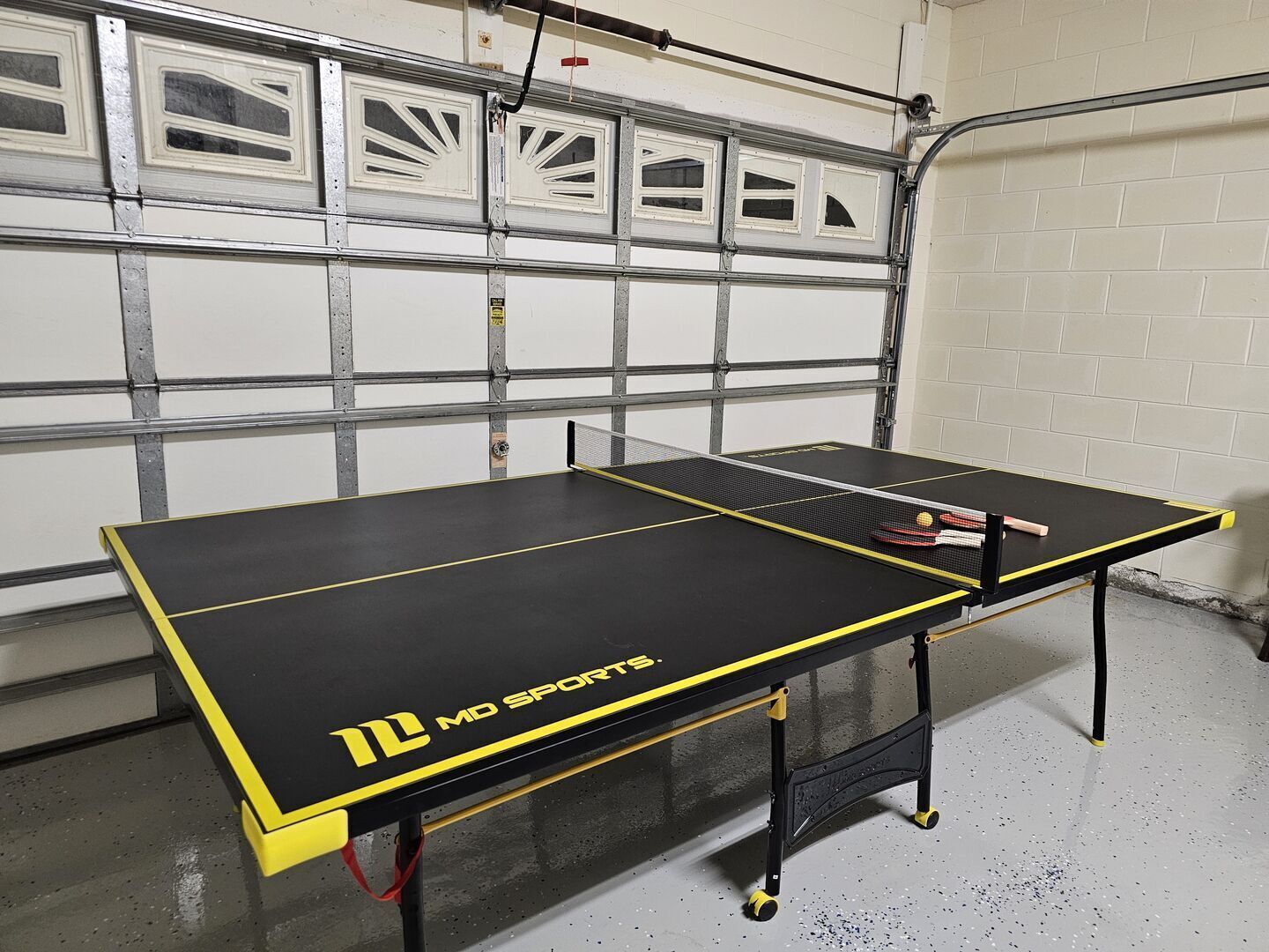 Ett bordtennisbord med MD Sports-logotypen, placerat i ett garage med en garageport och hyllor av metall.