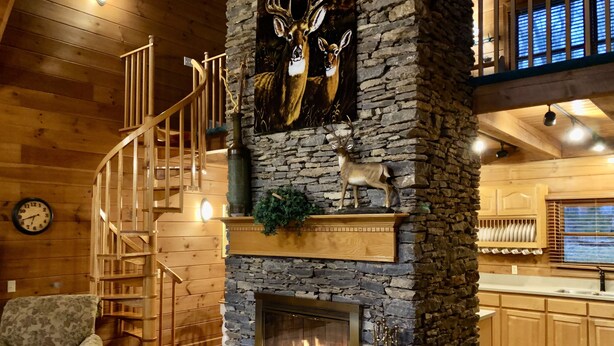 A cozy cabin interior with a stone fireplace, wooden staircase, and a deer wall art.