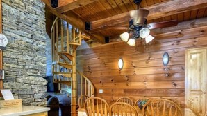A wooden interior with a stone wall, a spiral staircase, a dining table with chairs, and a ceiling fan.