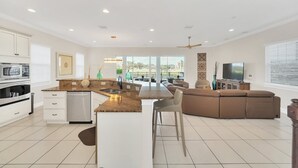 A modern kitchen with white cabinetry, a built-in oven, and a dishwasher. There is a dining area with a large window, a ceiling fan, and a flat-screen TV.