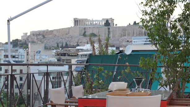 A terrace with outdoor seating, a view of ancient ruins in the background, and a modern building in the foreground. - Acropolis Home (Athens)