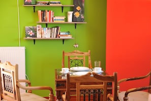 A dining area with a table set for two, surrounded by wooden chairs, against a green wall with shelves holding books and decorative items.