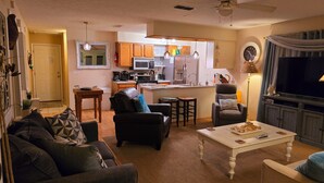 A living room with a sofa, a coffee table, and a TV. There is a kitchen in the background with cabinets and appliances. - A Special Getaway! (Panama City Beach)