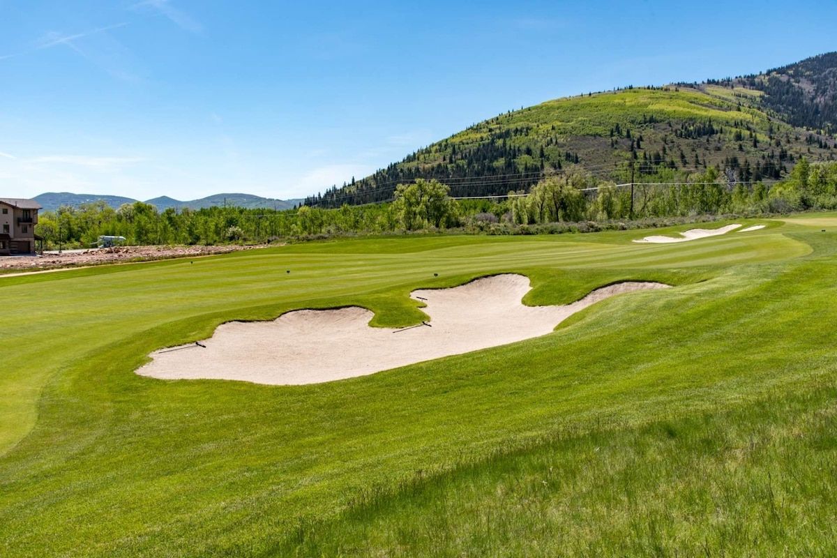 A golf course with bunkers, a clubhouse, and a hilly backdrop.