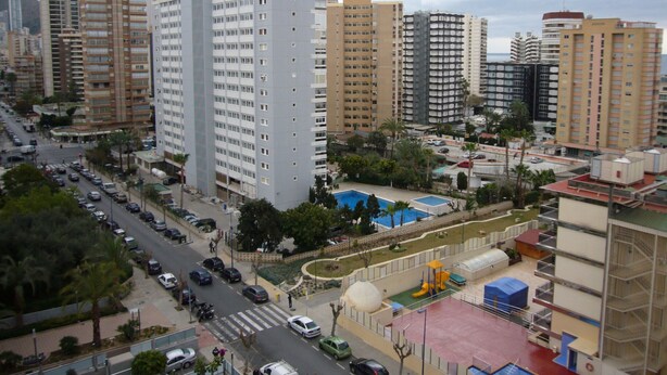 Un edificio alto con varios pisos, rodeado de otros edificios y un estacionamiento.