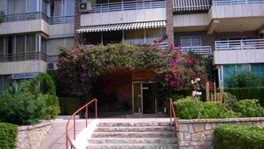 Entrada de un edificio con escalones de piedra y barandilla metálica, rodeada de follaje y un arco cubierto de flores.