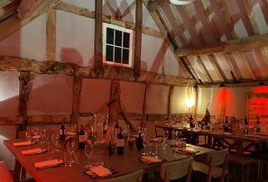 A rustic dining area with wooden beams, a small window, and tables set for a meal with wine glasses and bottles.