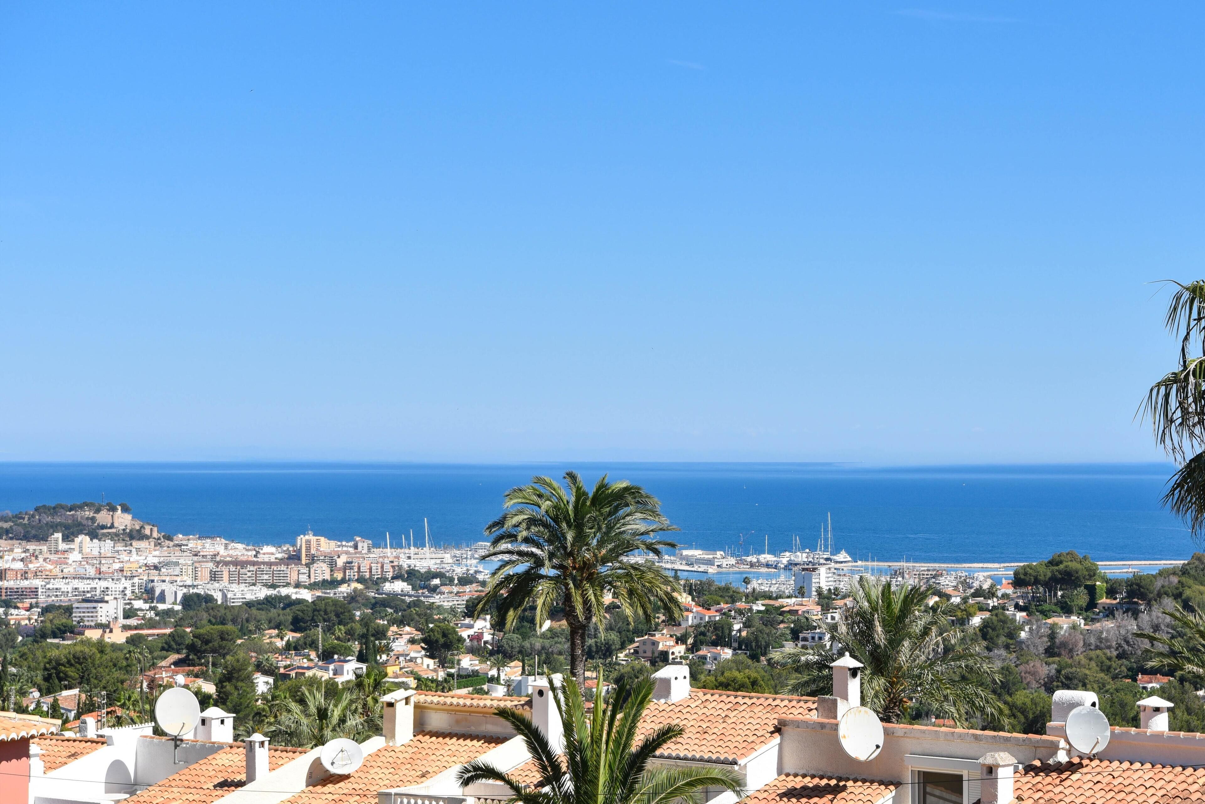 A coastal cityscape with a view of the sea, featuring residential buildings with terracotta roofs and satellite dishes.