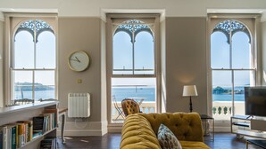 A living room with a yellow sofa, a bookshelf, a clock, and large windows with a view of the sea. - Luxurious 2 bed apartment with balcony and spectacular sea views (Plymouth)