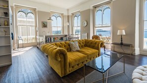 A spacious living room with a mustard sofa, a glass coffee table, and a view of the sea through large windows. - Luxurious 2 bed apartment with balcony and spectacular sea views (Plymouth)