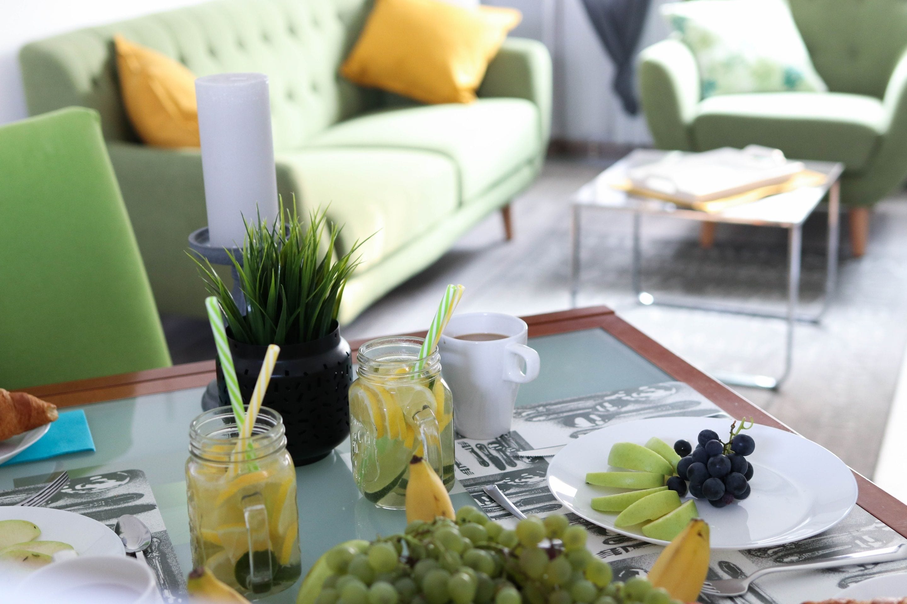 Een eettafel met een glazen blad, een witte mok, een bord met druiven en een banaan, en een glazen pot met een rietje.
