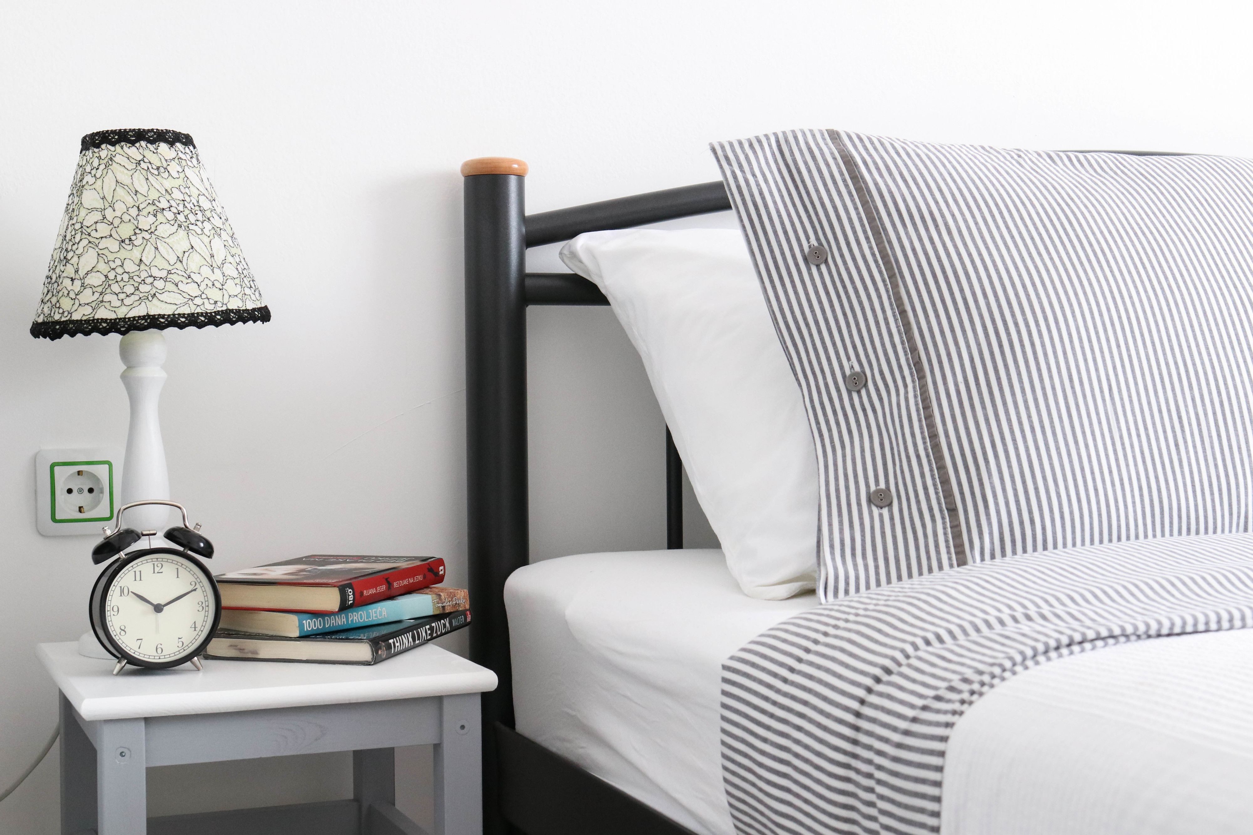 A neatly made bed with striped bedding, a bedside lamp with a lace shade, and a stack of books on a small bedside table.