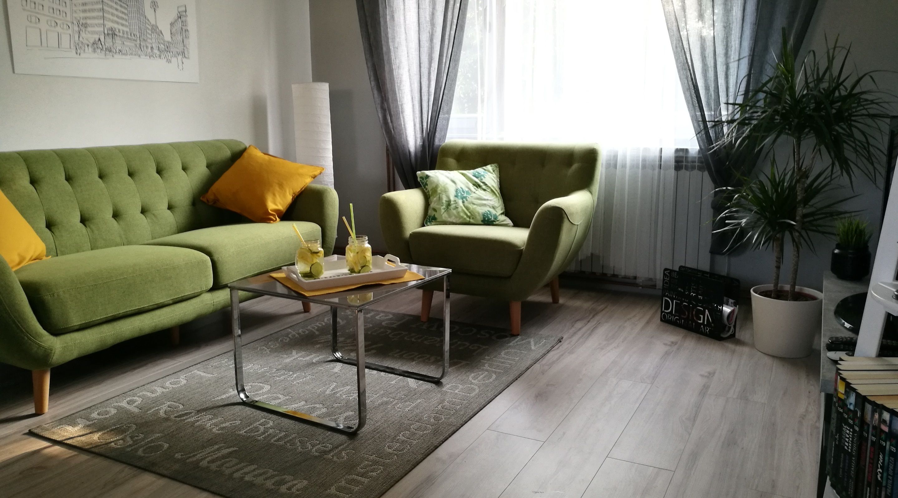 Een moderne woonkamer met een groene bank, een groene fauteuil en een salontafel met drankjes. Gordijnen, een potplant en een boekenplank zijn ook aanwezig.