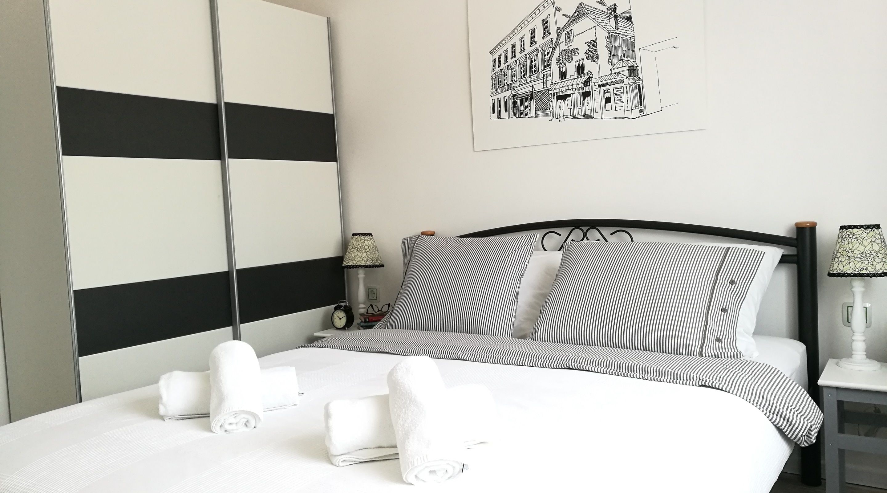 Un lit bien fait, avec une literie à rayures, une tête de lit noire et blanche à rayures, et une œuvre d’art représentant un bâtiment, accrochée au mur.