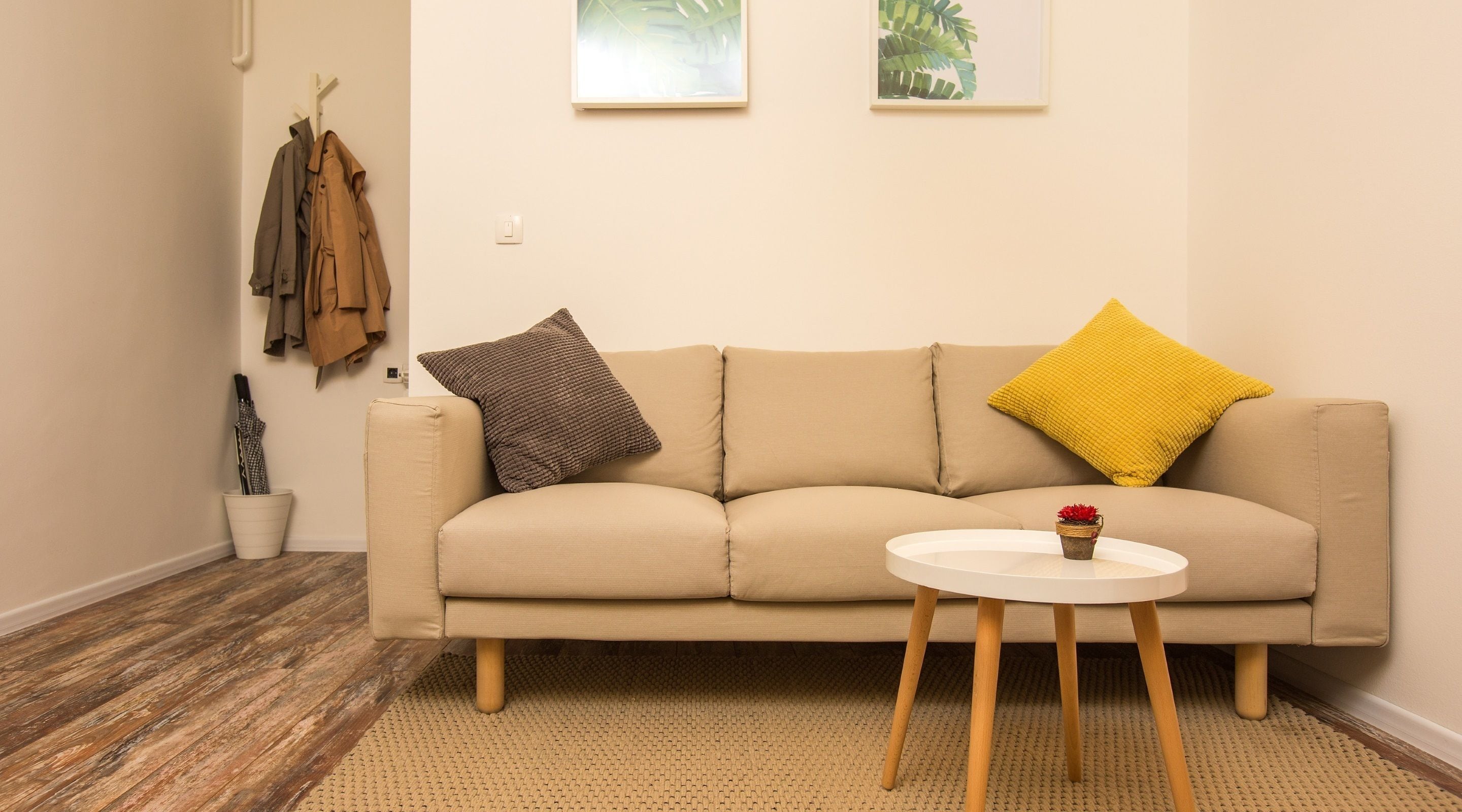 A beige sofa with two pillows, a small white table with a plant, and a wooden floor.