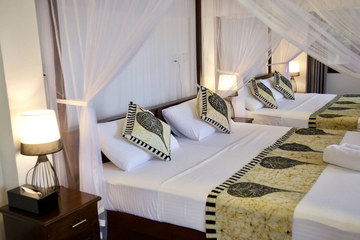 A hotel room with two beds, each with white bedding and patterned pillows. There is a nightstand with a lamp and a box, likely for toiletries.