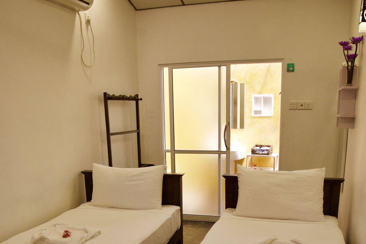 A hotel room with two single beds, each with a white pillow and a folded towel. There is a small table between the beds, and a wall-mounted air conditioner above the left bed.