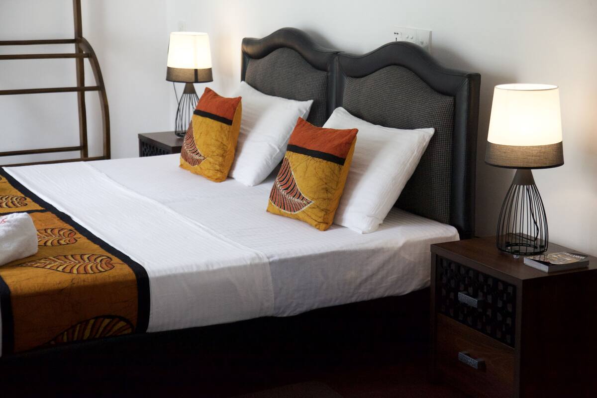 A neatly made bed with white linens and decorative pillows, a bedside lamp, and a nightstand with a book and a small decorative item.
