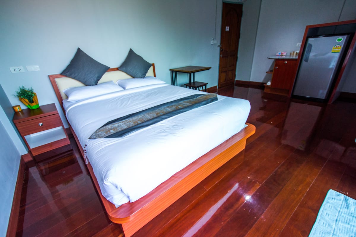 A neatly made bed with a wooden frame, a small bedside table with a plant, and a refrigerator in the corner.
