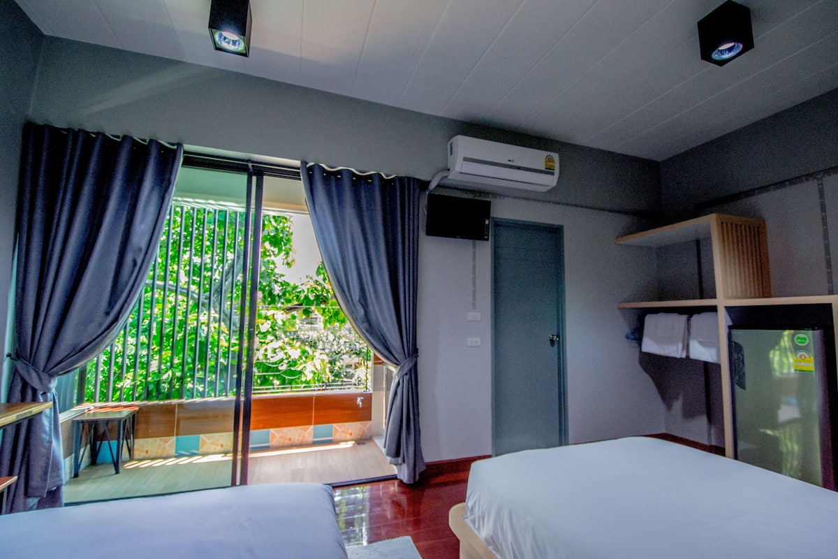 A hotel room with two beds, a balcony with curtains, an air conditioning unit, a door, and a small table.