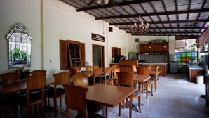 A dining area with wooden tables and chairs, a kitchen in the background, and a ceiling fan. - Griya Gendhis Saraswati (Klaten)