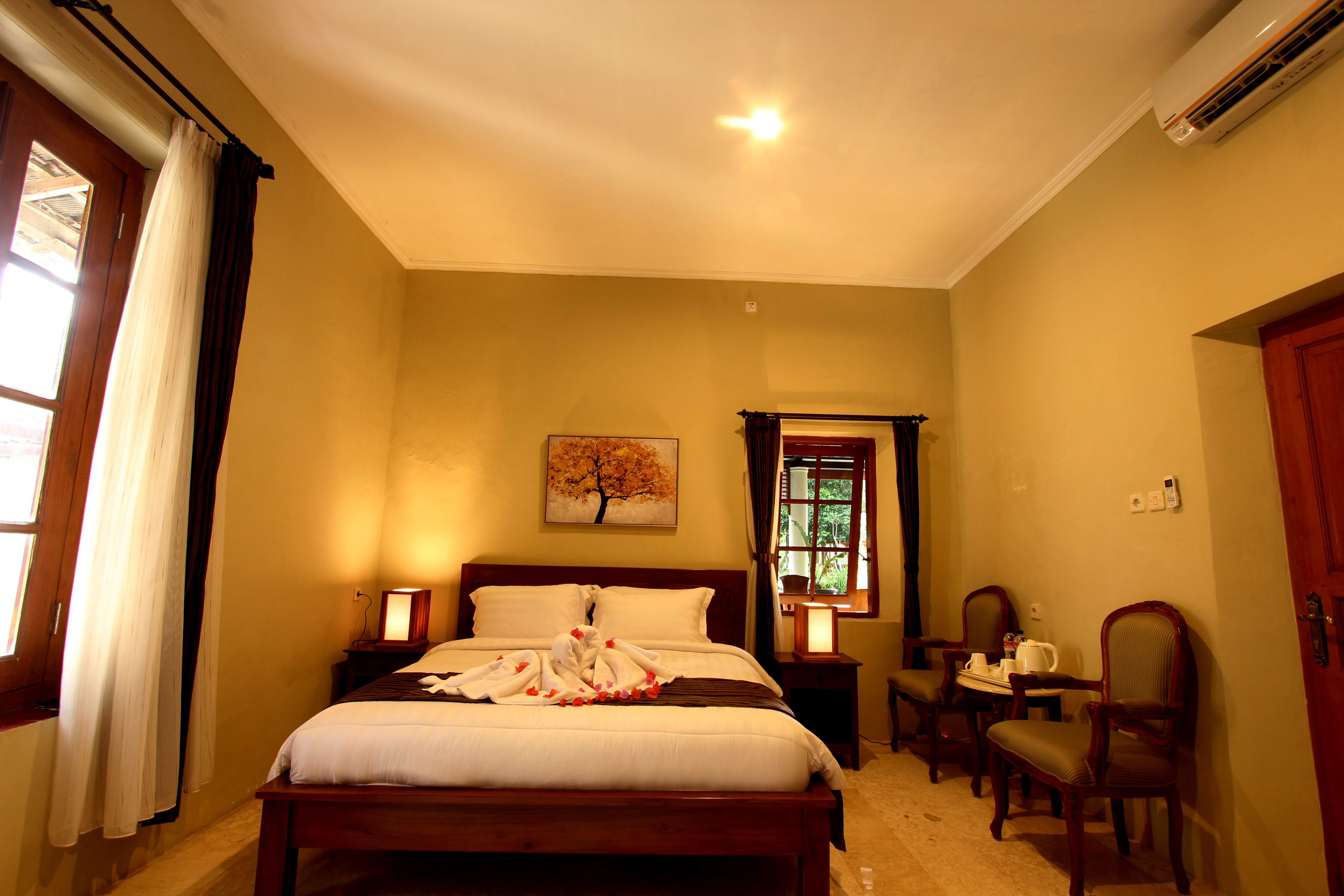 A hotel bedroom with a bed, bedside tables, a desk with chairs, a window, and a painting on the wall.
