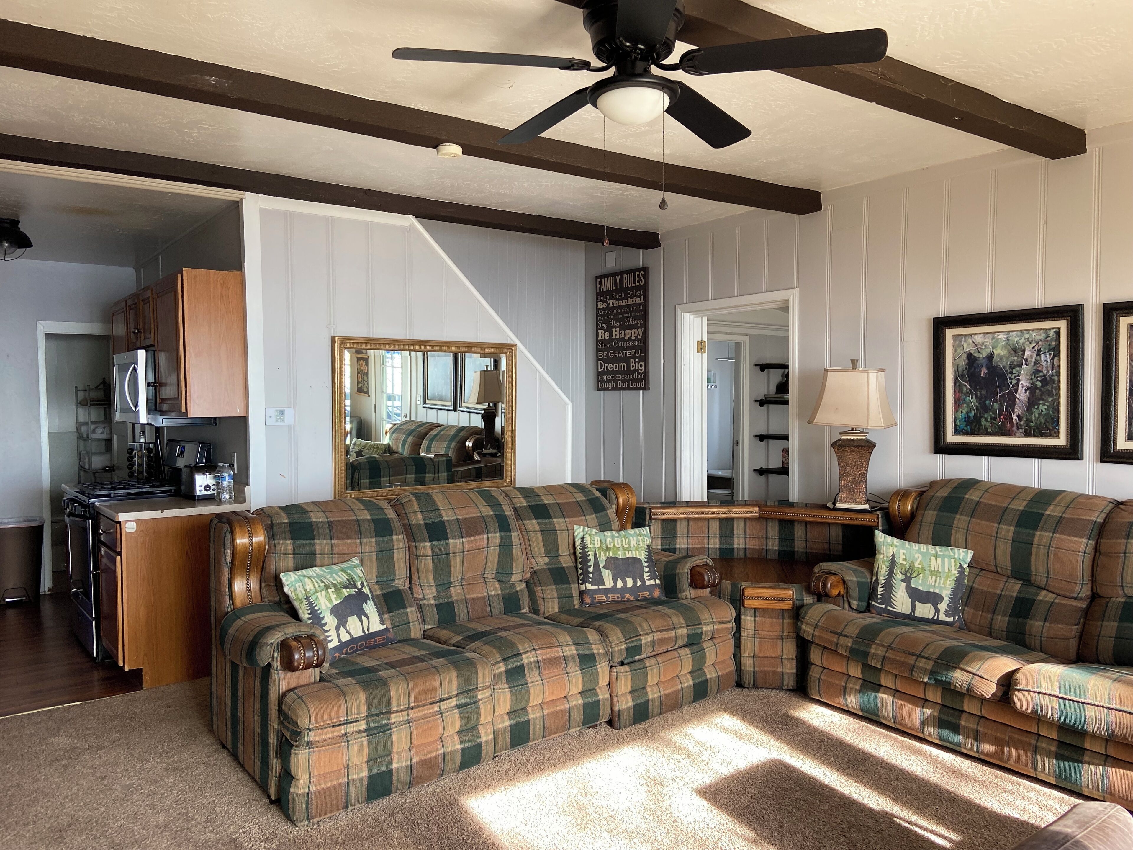 A cozy living room with a fireplace, plaid sofas, a ceiling fan, and a kitchen area.