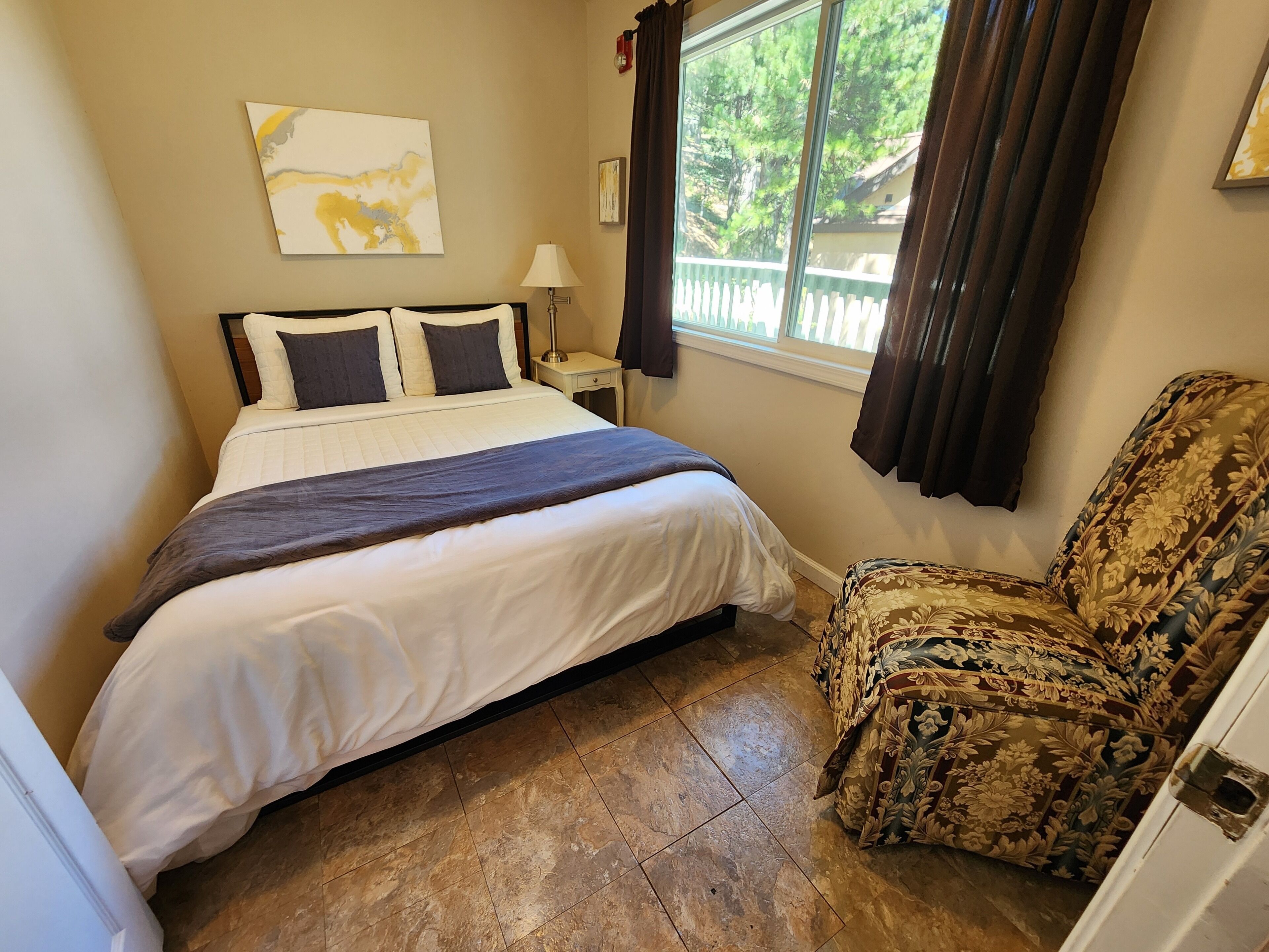 A bedroom with a bed, a chair, a window with curtains, a lamp, and a wall art piece.